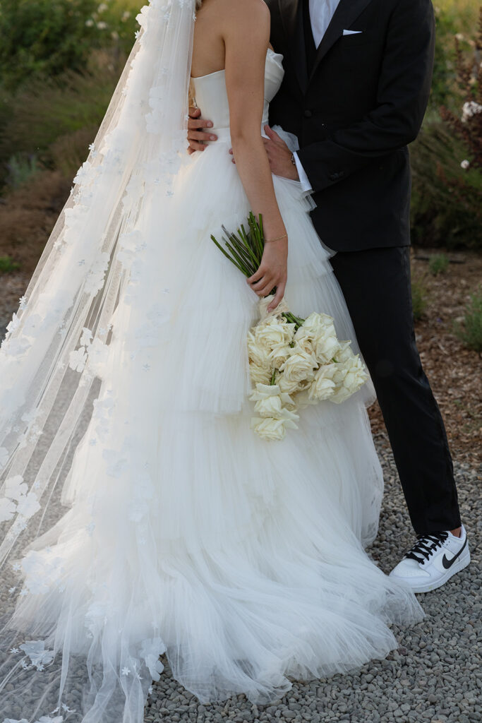 Bride and groom embrace outdoors; bride in white gown holds white roses, groom in black suit with sneakers.