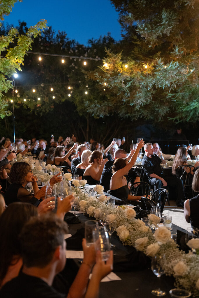 Guests seated at a long table raise glasses for a toast during an outdoor evening event, with string lights and floral decorations.
