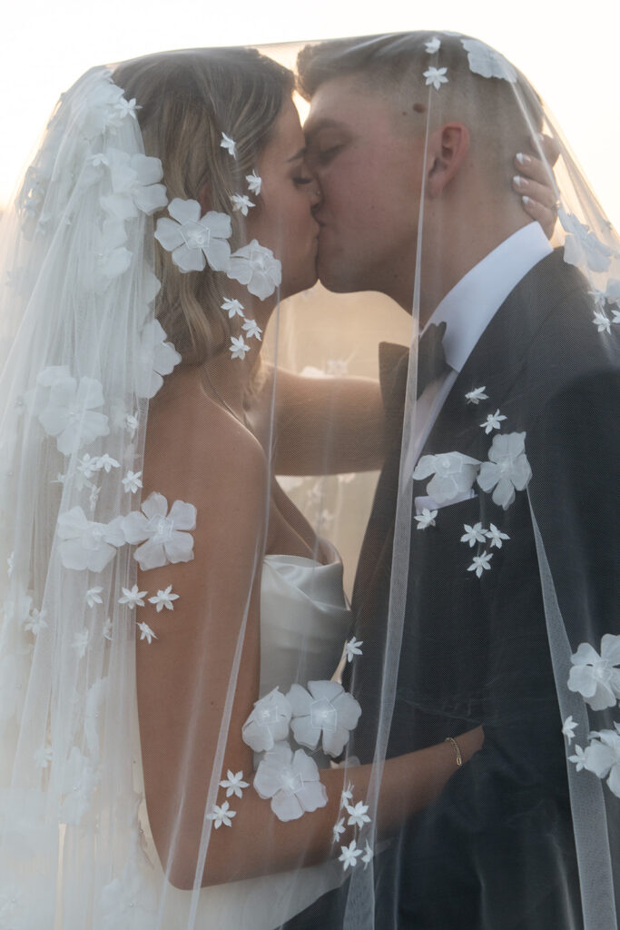 A bride and groom kiss under a veil adorned with floral designs during their wedding.