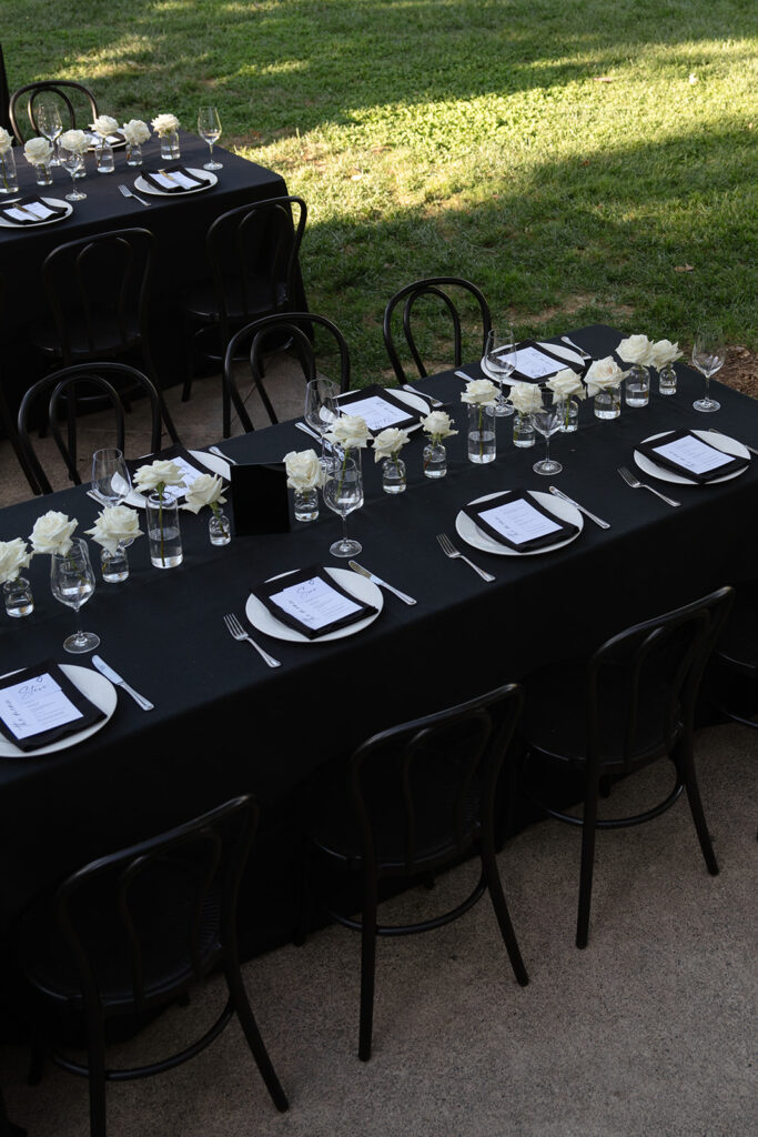 Two elegantly set dining tables with black tablecloths, white flowers, plates, and glasses on a grassy outdoor setting.