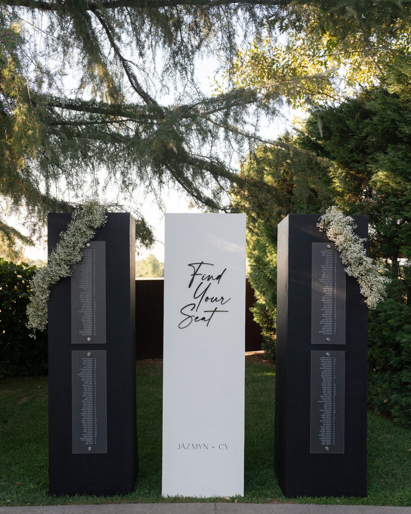 A seating chart display outdoors, featuring a central white sign reading "Find Your Seat" flanked by black columns adorned with floral arrangements and guest names.
