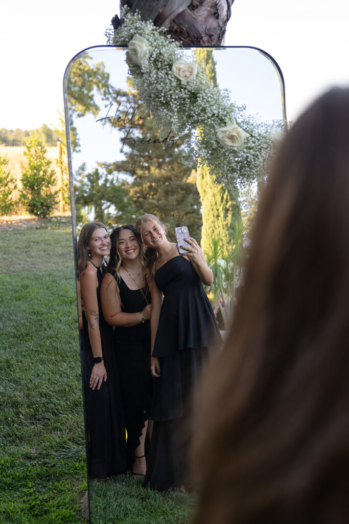 Three women in black dresses smile while taking a selfie in a decorative mirror outdoors.