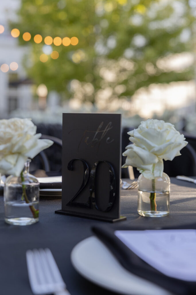 Outdoor table setting with a "Table 20" sign between two white roses in glass vases. Background features blurred trees and string lights.