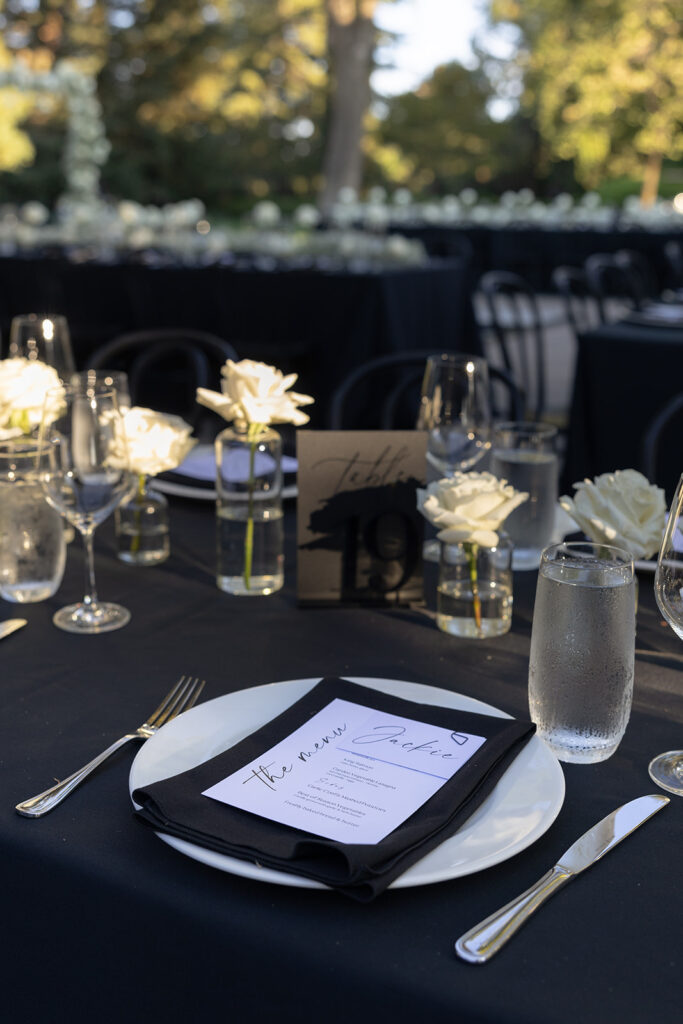 Elegantly set table with black tablecloth, menus, white roses in glass vases, and glassware at an outdoor event.