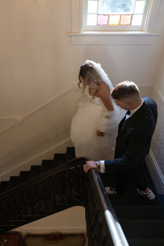 A bride and groom ascend a staircase with a stained glass window above them. The groom is dressed in a suit, and the bride is in a white gown.