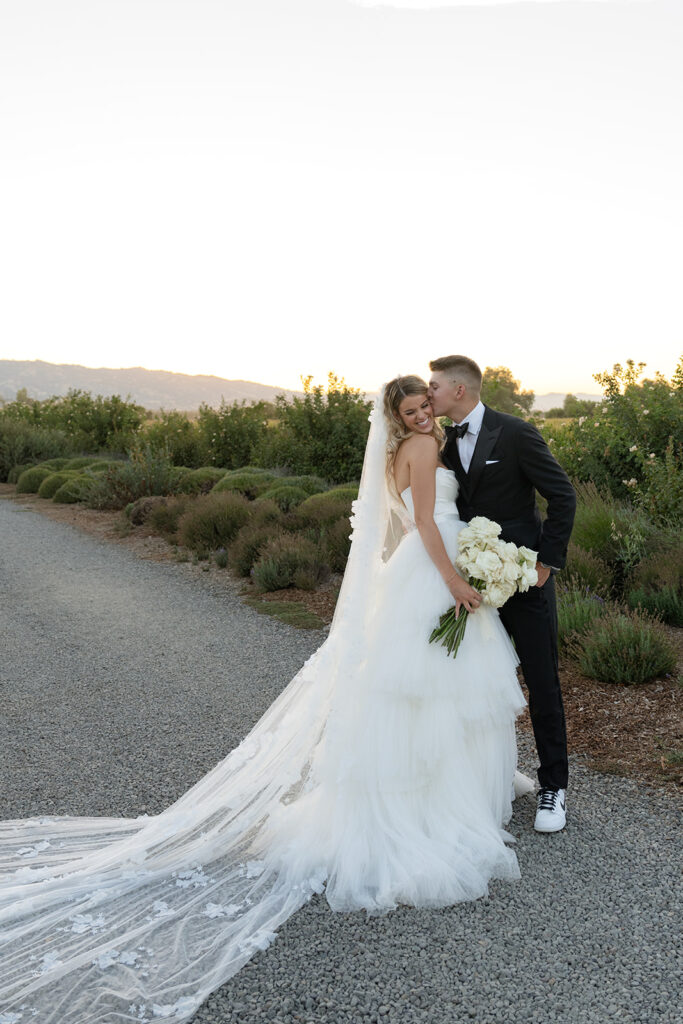 A bride and groom stand on a gravel path, with the groom kissing the bride's cheek. She holds a bouquet of white flowers, and they are surrounded by greenery at sunset.