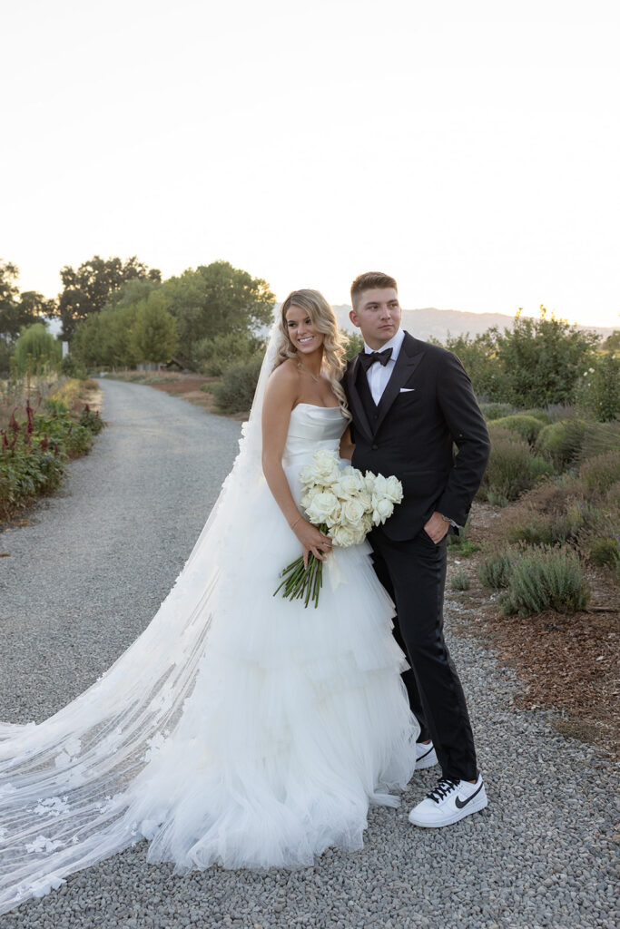 A bride in a white gown and a groom in a black suit pose on a gravel path, holding a bouquet of white flowers, with greenery and hills in the background.