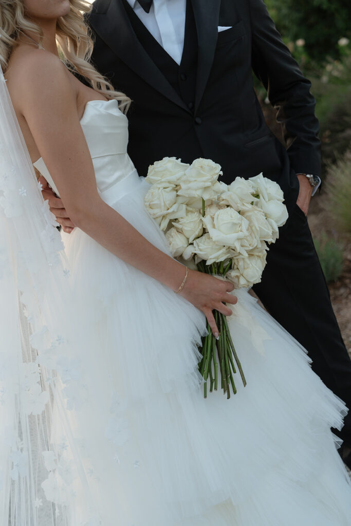 Bride in a white gown holds a bouquet of white roses, standing next to a man in a black suit, outdoors.