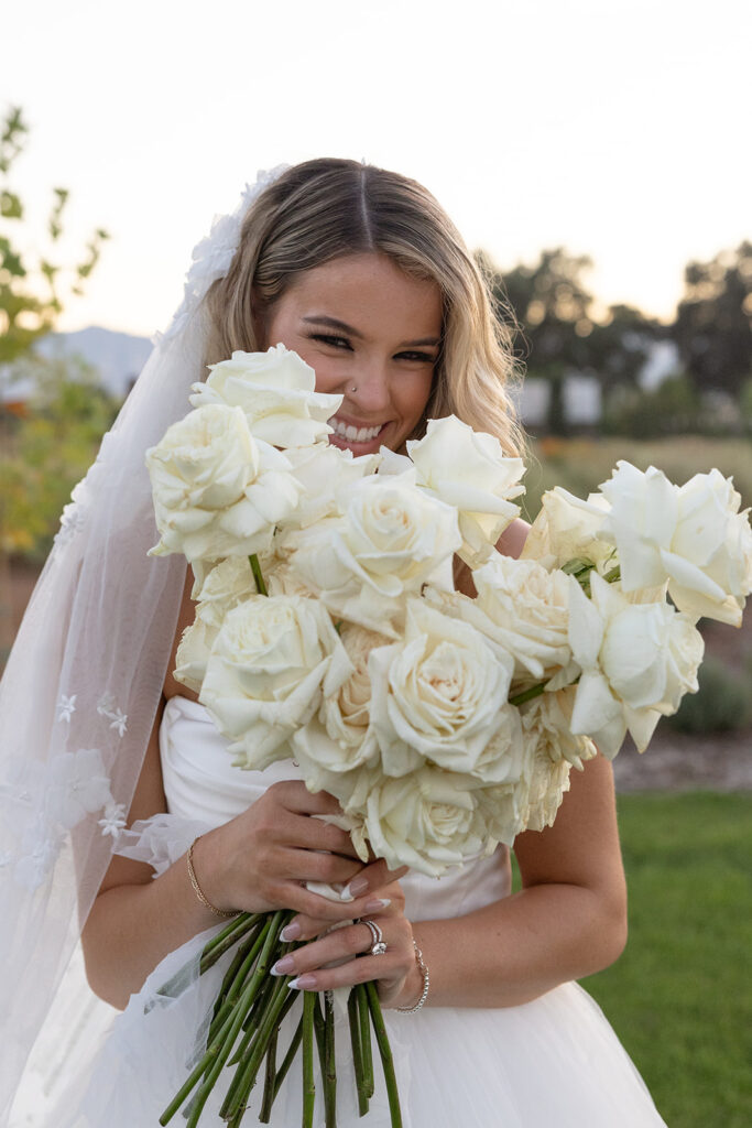 A bride in a white dress holds a bouquet of white roses, smiling outdoors.