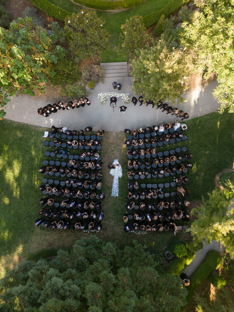 Aerial view of an outdoor wedding ceremony with guests seated in rows, a couple standing at the altar, surrounded by greenery and pathways.