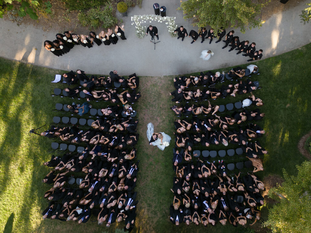 Aerial view of an outdoor wedding ceremony with guests seated in rows on either side of the aisle, where a couple stands in front of an officiant.