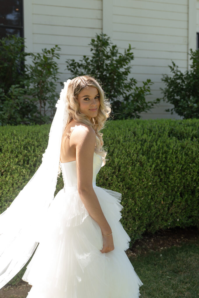 A bride in a white wedding dress and veil stands outdoors in front of green hedges and a white building, looking back over her shoulder.