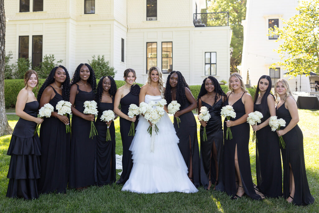 A bride in a white dress stands with eleven bridesmaids in black dresses, all holding white bouquets, in front of a large white building.