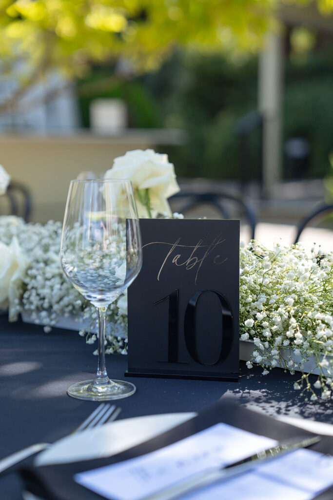A table setting features a "Table 10" sign, a wine glass, and a centerpiece of white flowers on a black tablecloth.
