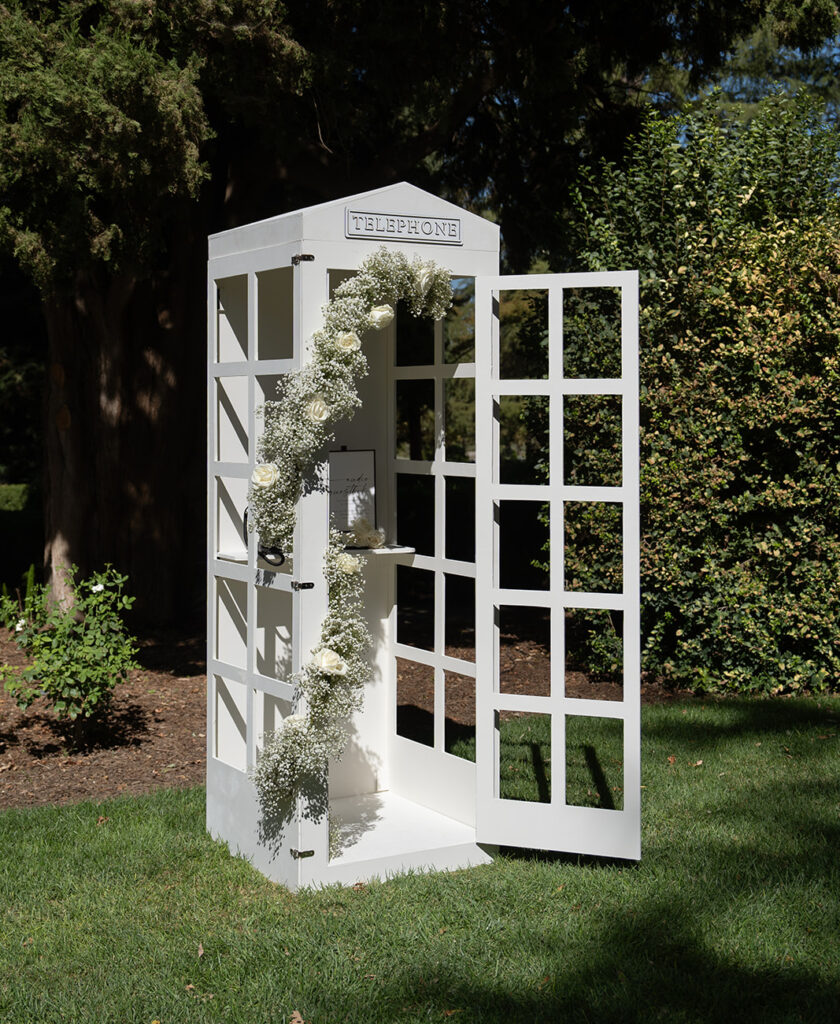 White telephone booth decorated with white flowers, located on a grassy area surrounded by trees.