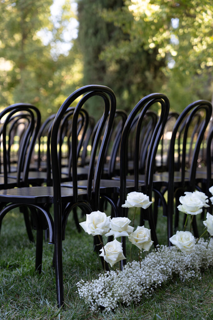 Rows of black chairs lined up outdoors on grass, with white roses and baby's breath flowers placed along the aisle.