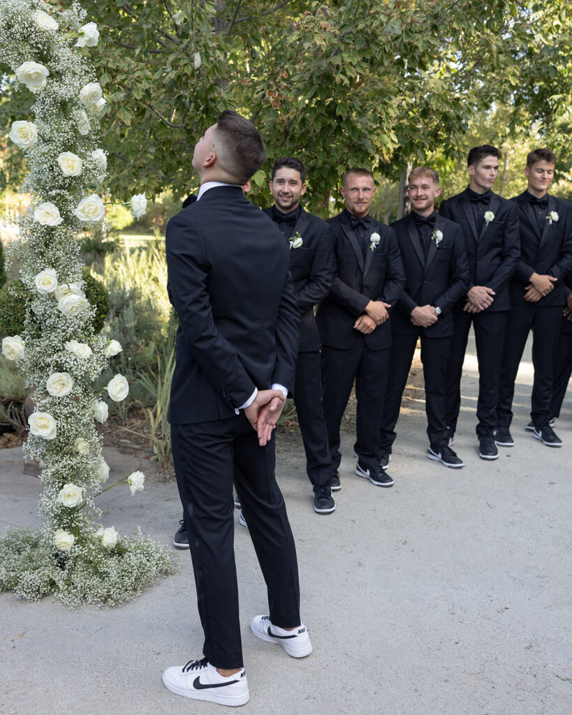 A groom in a black suit and white sneakers stands under a floral arch, looking up. Five groomsmen in matching outfits and holding white roses stand nearby, smiling.