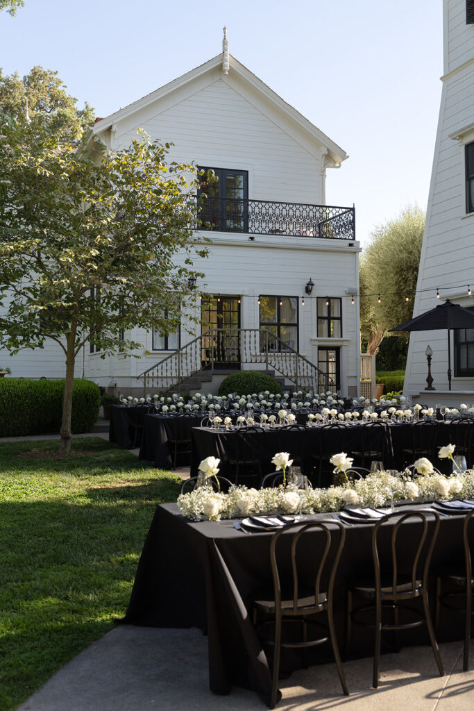 Outdoor wedding reception setup with black tablecloths, white floral centerpieces, and black chairs, next to a white house with a balcony and a tree.
