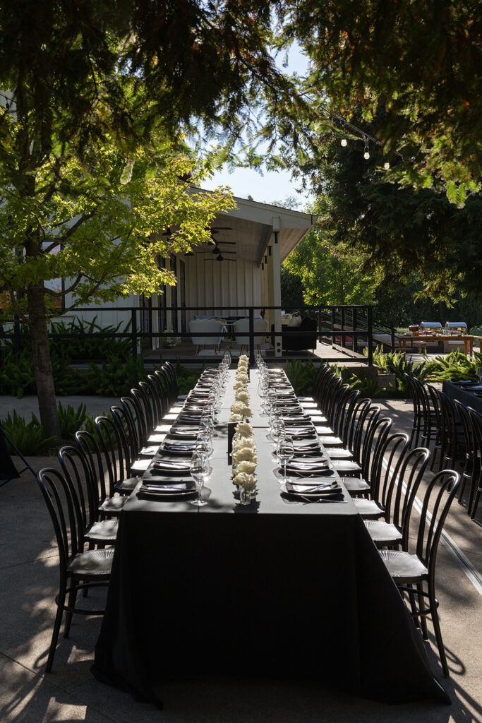Outdoor dining setup with a long table, black chairs, and table settings including white flowers, plates, and glasses under a shady tree canopy.