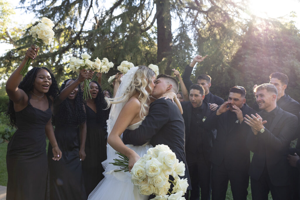 A bride and groom kiss outdoors, surrounded by bridesmaids in black dresses holding flowers and groomsmen in black suits, with trees and sunlight in the background.