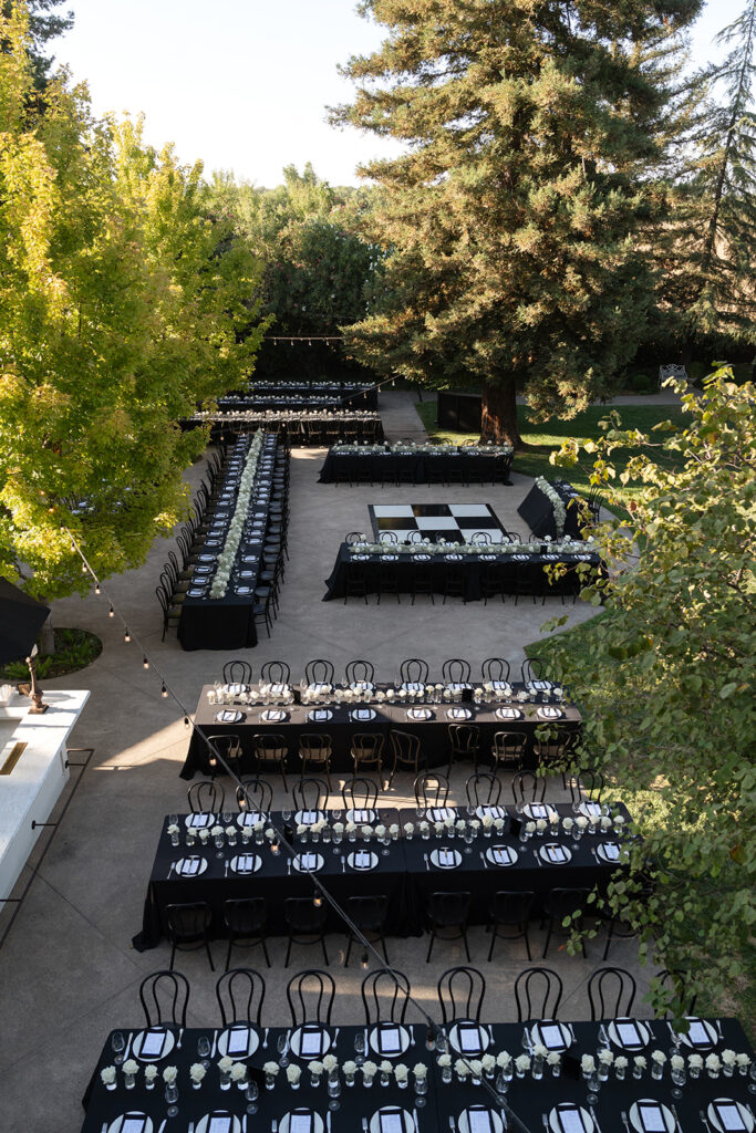 Outdoor event setup with long tables covered in black tablecloths and arranged in a U-shape under string lights, surrounded by trees.