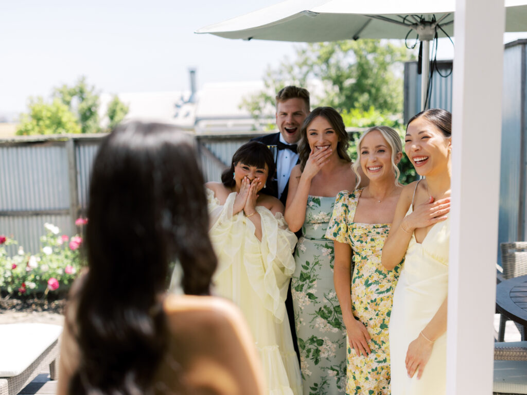 A group of five people in formal attire stand under an umbrella, smiling and reacting joyfully towards a person in the foreground.
