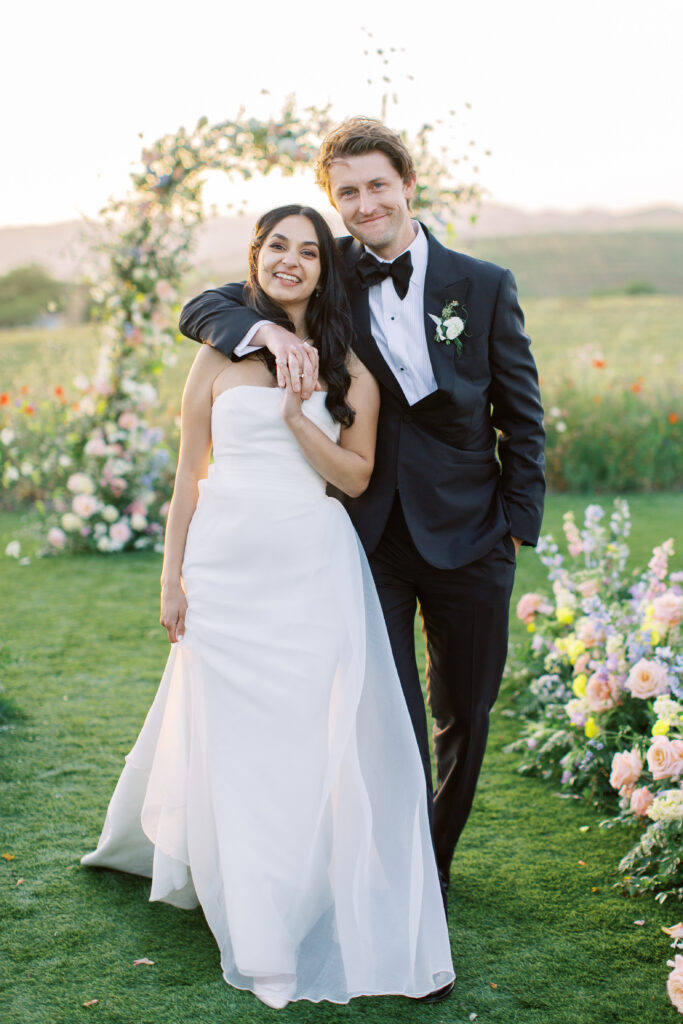 A bride and groom stand together outdoors in formal wedding attire, with a floral arch and scenic landscape in the background.