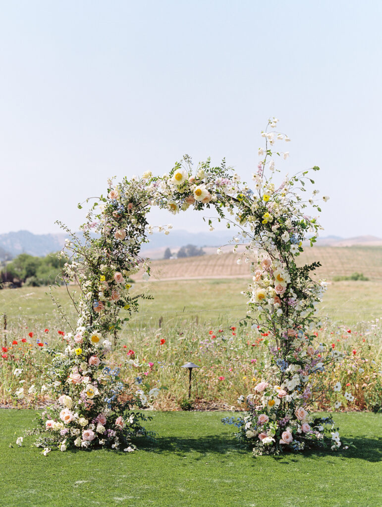 A floral arch made of pastel flowers stands on a grassy field with a backdrop of rolling hills and flowers.