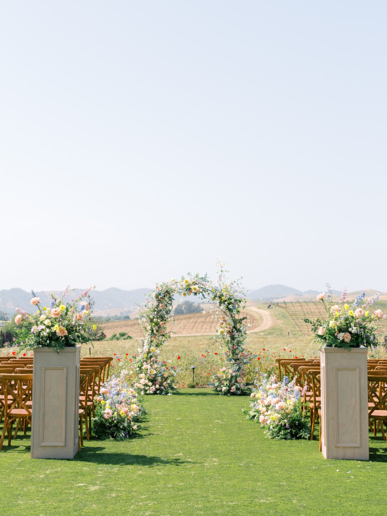Outdoor wedding setup with a floral arch, wooden chairs, and decorative flower arrangements on a green lawn, with a scenic landscape in the background.