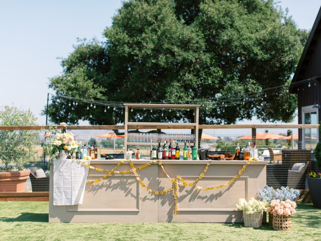 Outdoor bar setup with various bottles and decorations, including a garland and flowers. Large tree in the background, sunny weather.