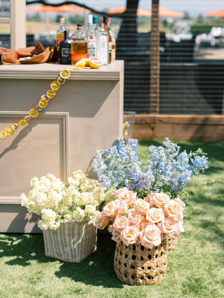 Bar setup outdoors with bottles and citrus garnish on a counter. Baskets of pink roses, white flowers, and blue flowers are placed on the grass below.
