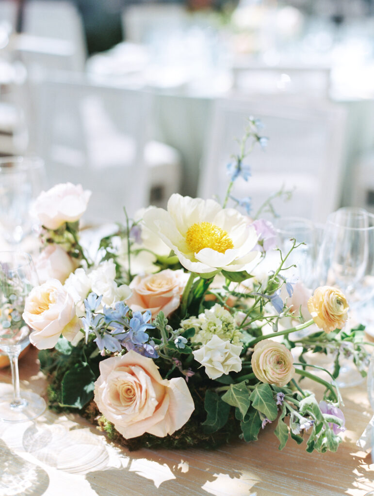 Floral centerpiece with white and pink flowers, including a prominent white peony and scattered greenery, on a table with glassware in the background.