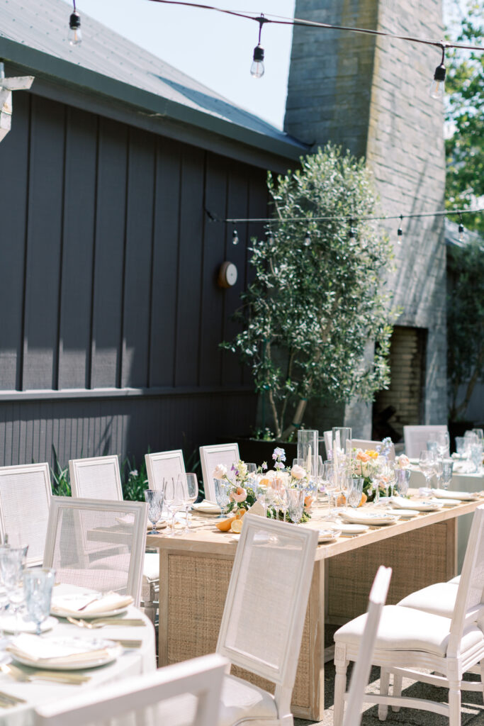 Outdoor dining setup with beige chairs around wooden tables. The tables are decorated with flowers and candles. A gray building and string lights are in the background.