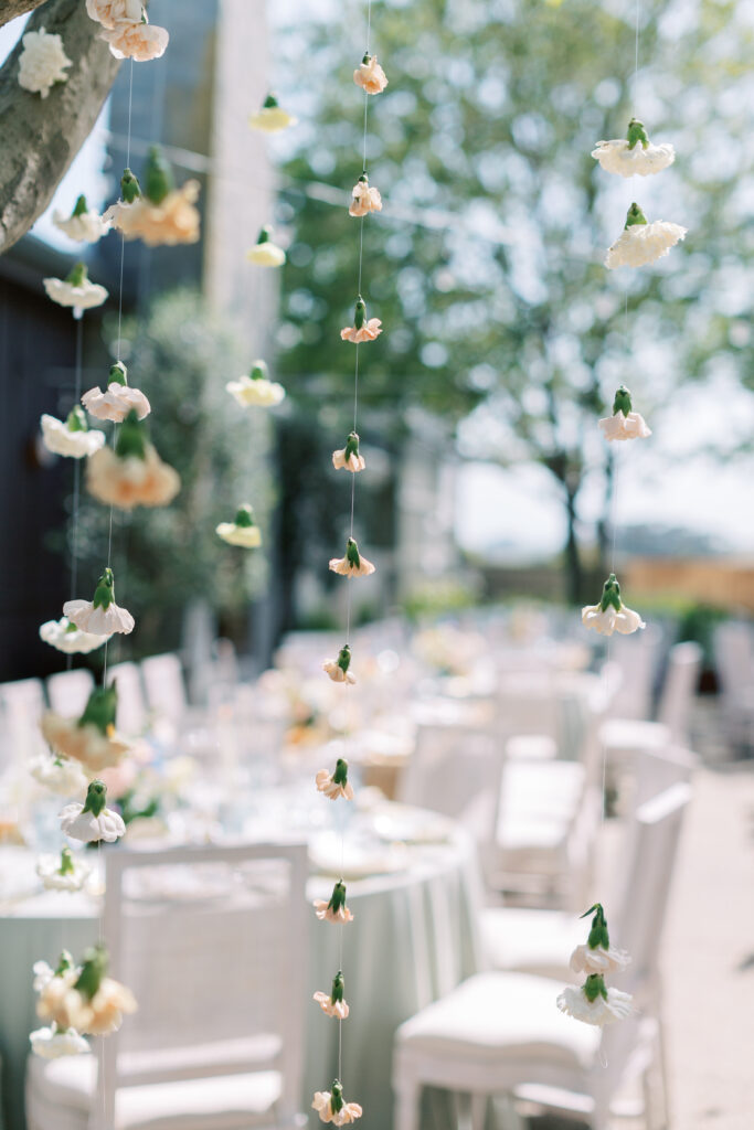 Hanging white and peach flowers on thin strings in focus; blurred outdoor setting with white chairs and tables in the background.