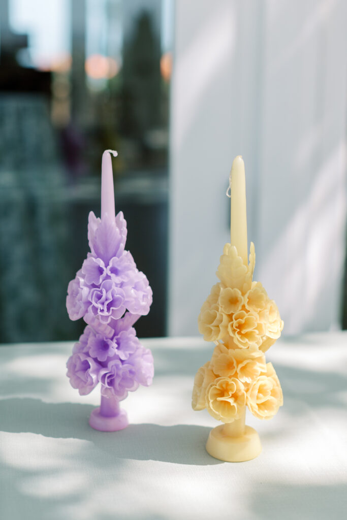 Two ornate candles, one purple and one yellow, with floral designs, stand on a table in sunlight.