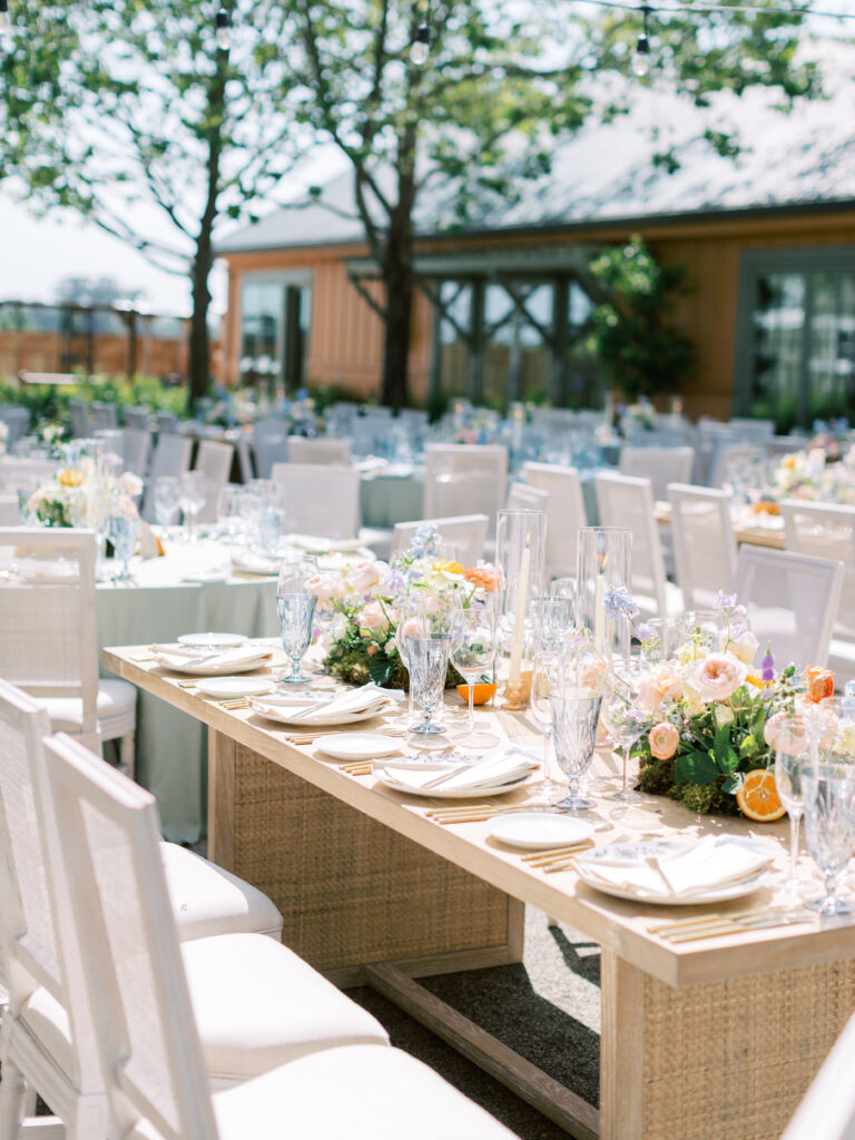 Outdoor wedding reception with elegantly set tables, decorated with floral centerpieces and glassware, arranged in a garden setting.