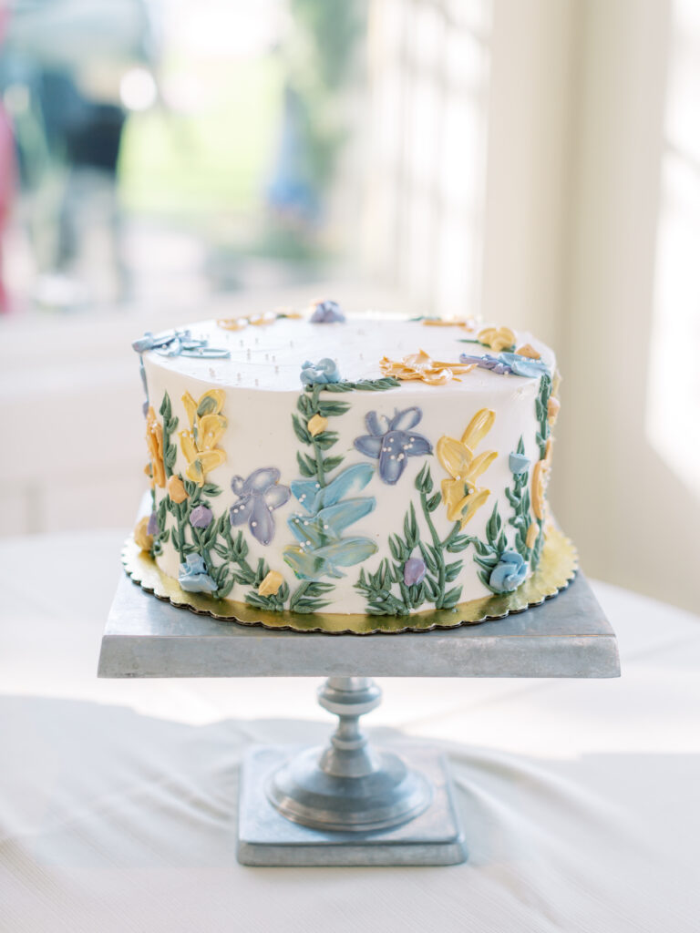 A round cake on a stand, decorated with colorful floral designs in icing.