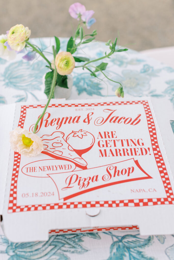 Pizza box with text: "Reyna & Jacob are getting married! The Newlywed Pizza Shop, 05.18.2024, Napa, CA," decorated with flowers and a floral tablecloth.