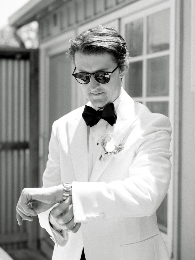 A man in a white tuxedo and sunglasses checks his watch outside near a building.
