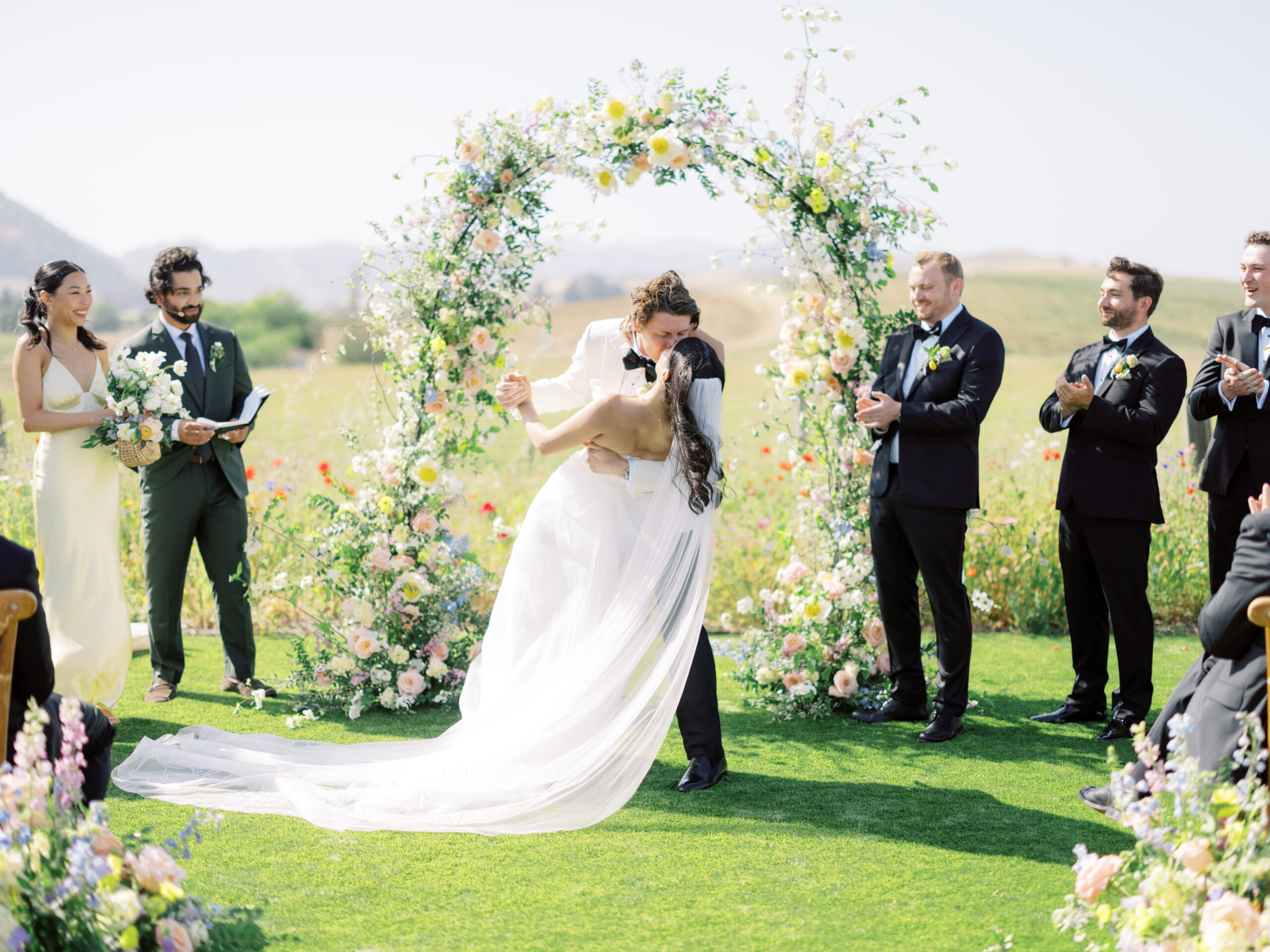 A couple kisses under a floral arch at an outdoor ceremony, flawlessly orchestrated by a Napa wedding planner, as the wedding party applauds.