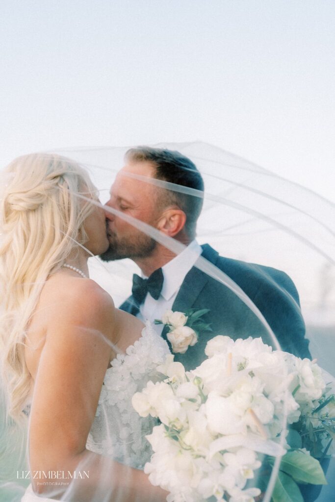 A bride and groom are kissing under a veil, holding a bouquet of white flowers.