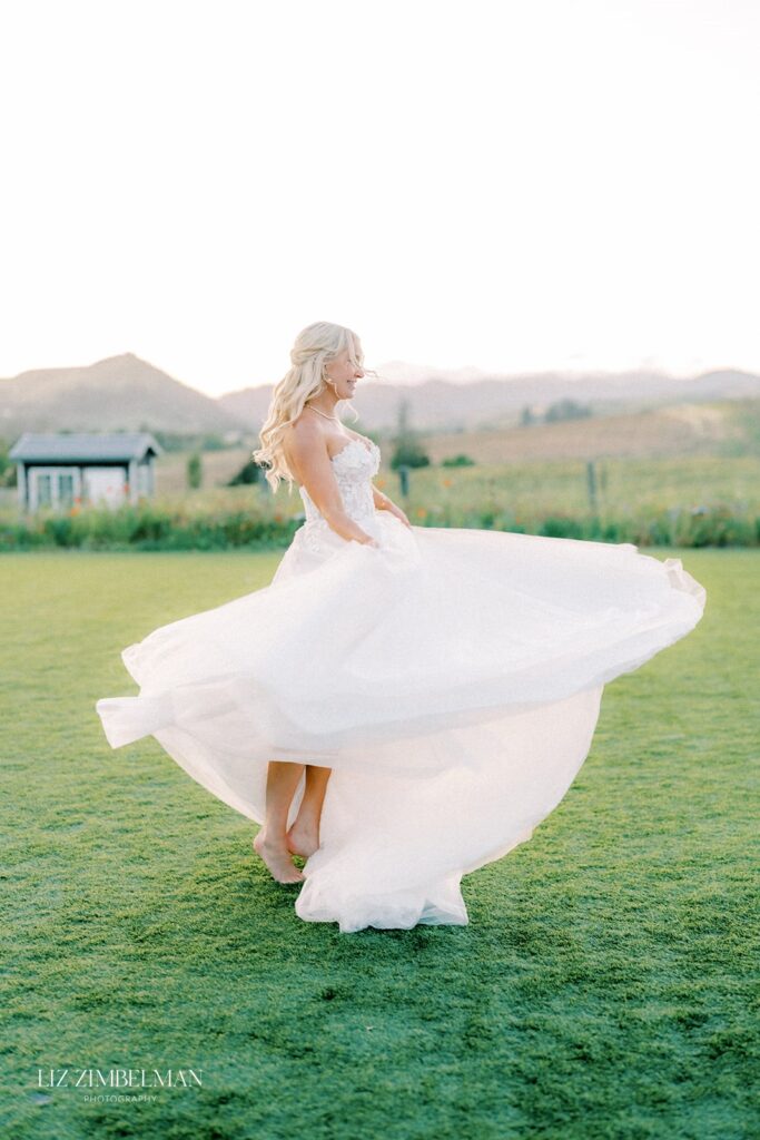 A bride in a white dress twirls on a grassy field with mountains and a small building in the background.