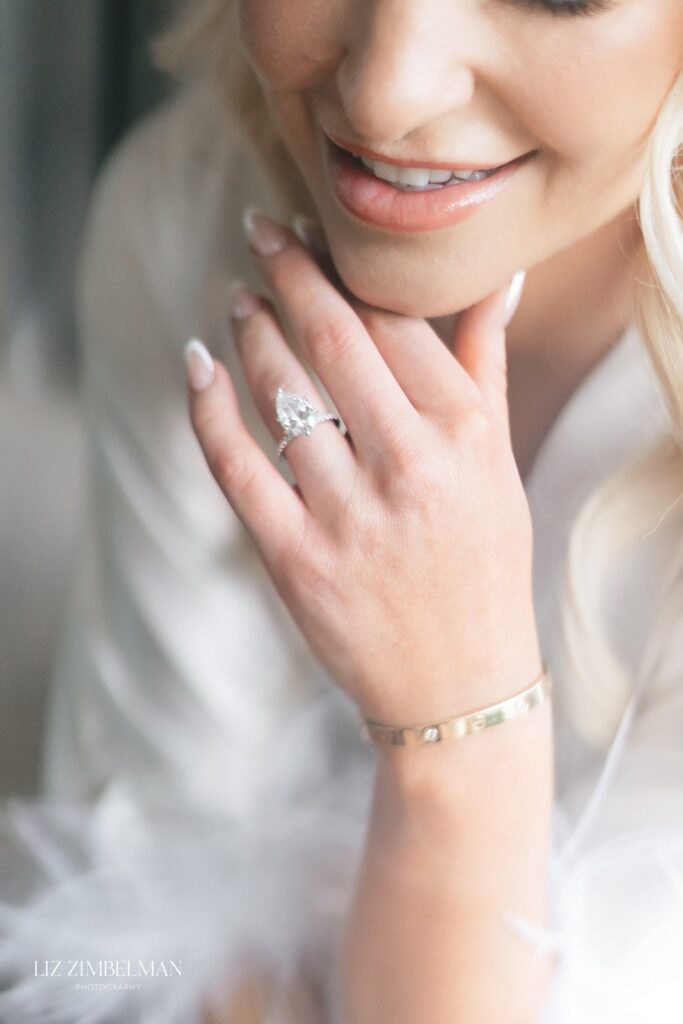 A person with long, blonde hair smiles while resting their chin on their hand, displaying a ring and a bracelet.