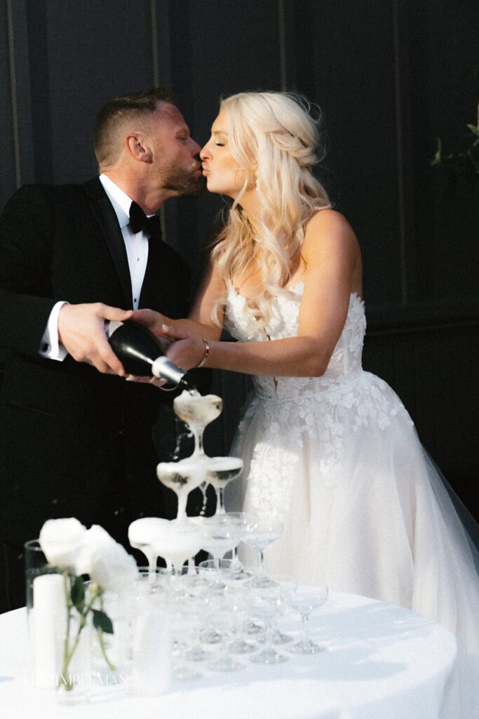 A couple in formal attire kisses while pouring champagne into a tower of glasses.