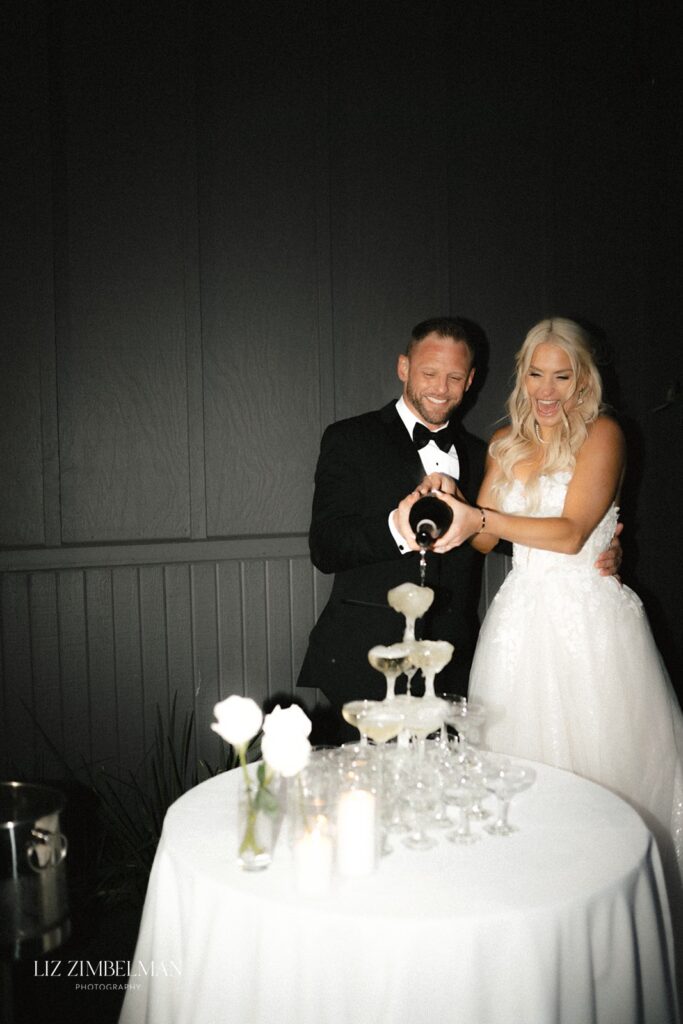 A couple in formal attire is pouring champagne into a tower of glasses on a table with candles and white roses.