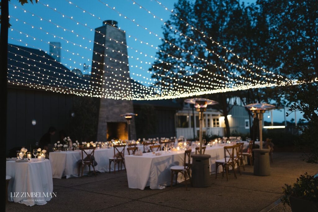 Sophia and Austin's wedding features an enchanting outdoor dining area at dusk, where long tables are elegantly set for the occasion. Overhead string lights cast a warm glow, while heaters keep guests cozy amidst the trees in the background.