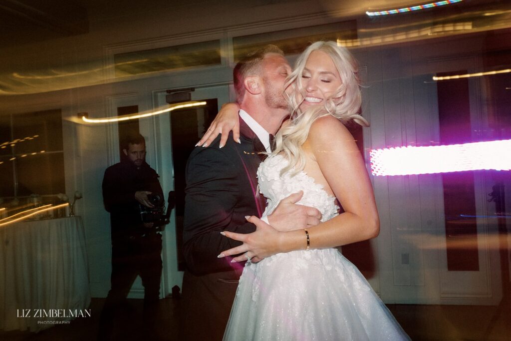 A couple dances closely, smiling, as colorful lights blur in the background. A person with a camera is visible in the background.