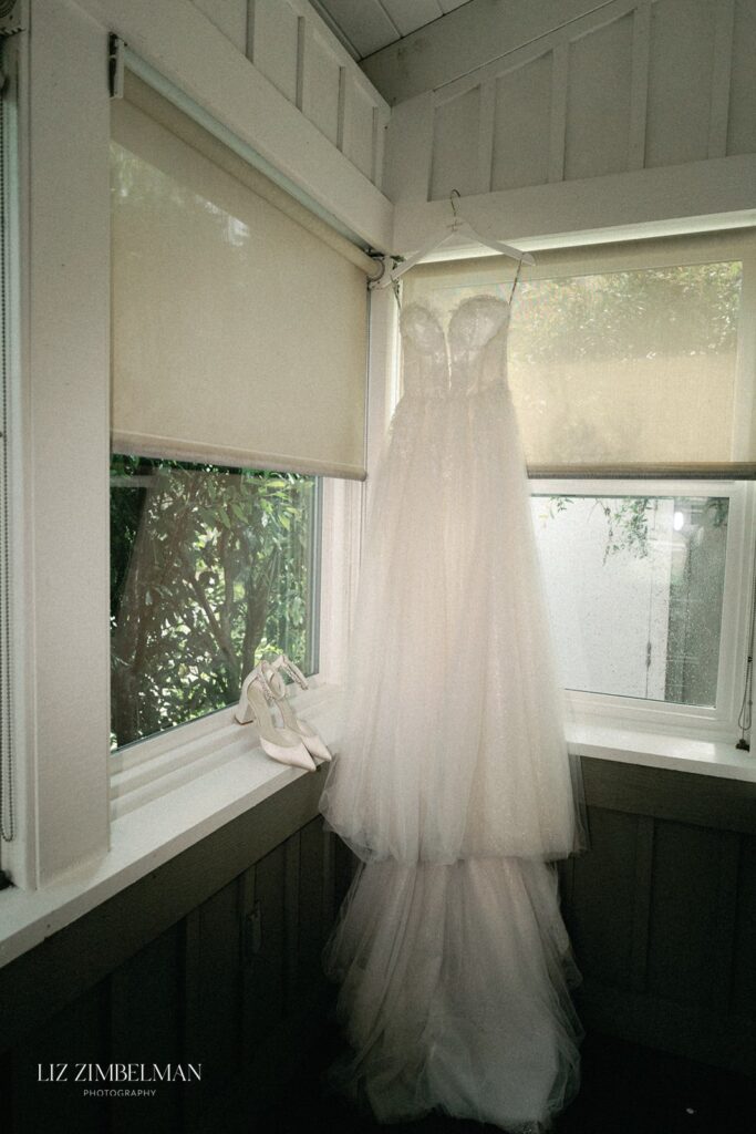 A white wedding dress hangs on a window frame beside a pair of white heels in a sunlit room.