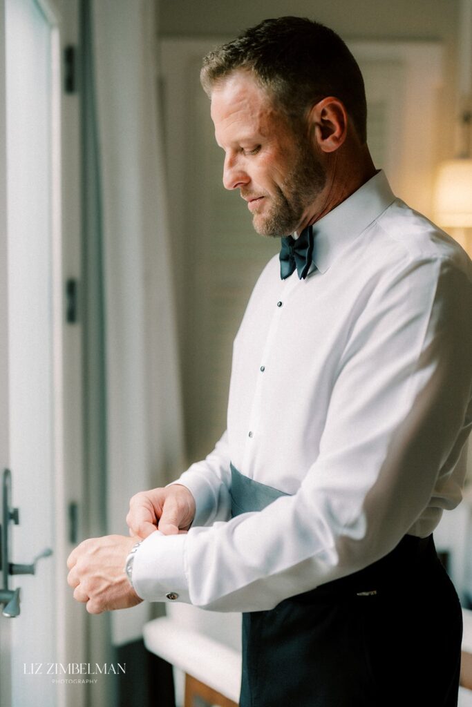 A man in formal attire adjusts his cufflink near a window, wearing a white shirt with a black bow tie and cummerbund.