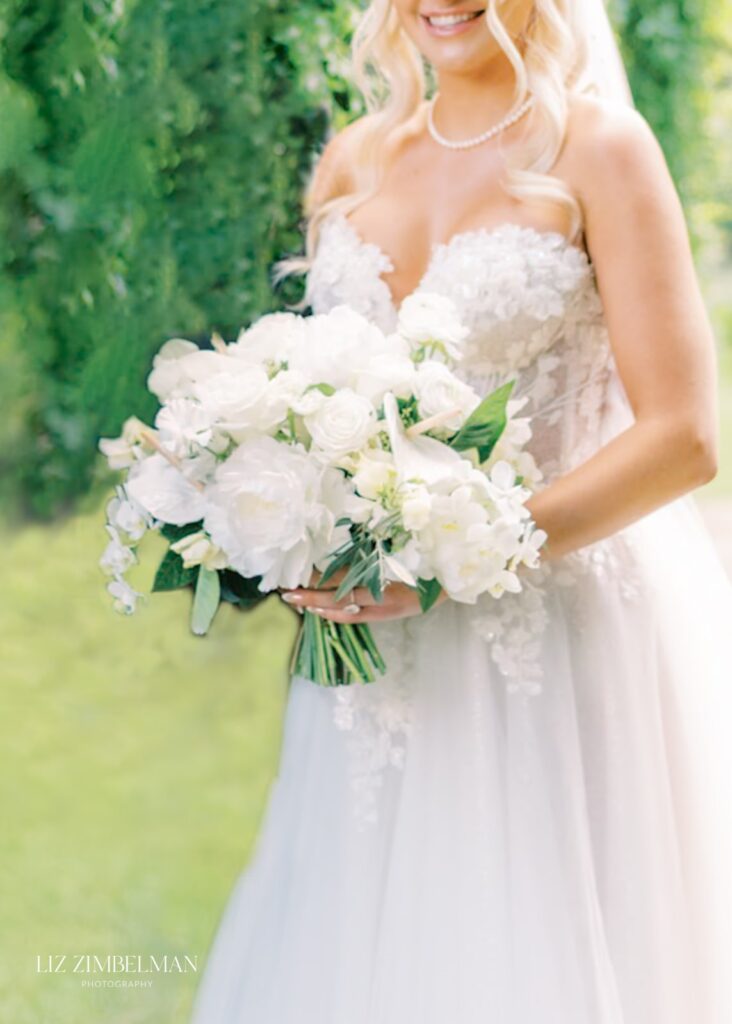 Bride in a white lace gown holding a bouquet of white flowers, standing outdoors with greenery in the background.
